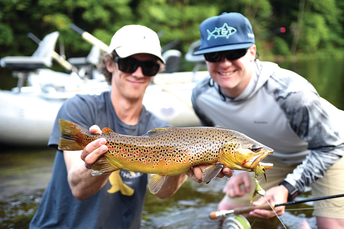 two anglers, one holding a large brown trout with two hands out of the water, the other squatting and smiling behind
