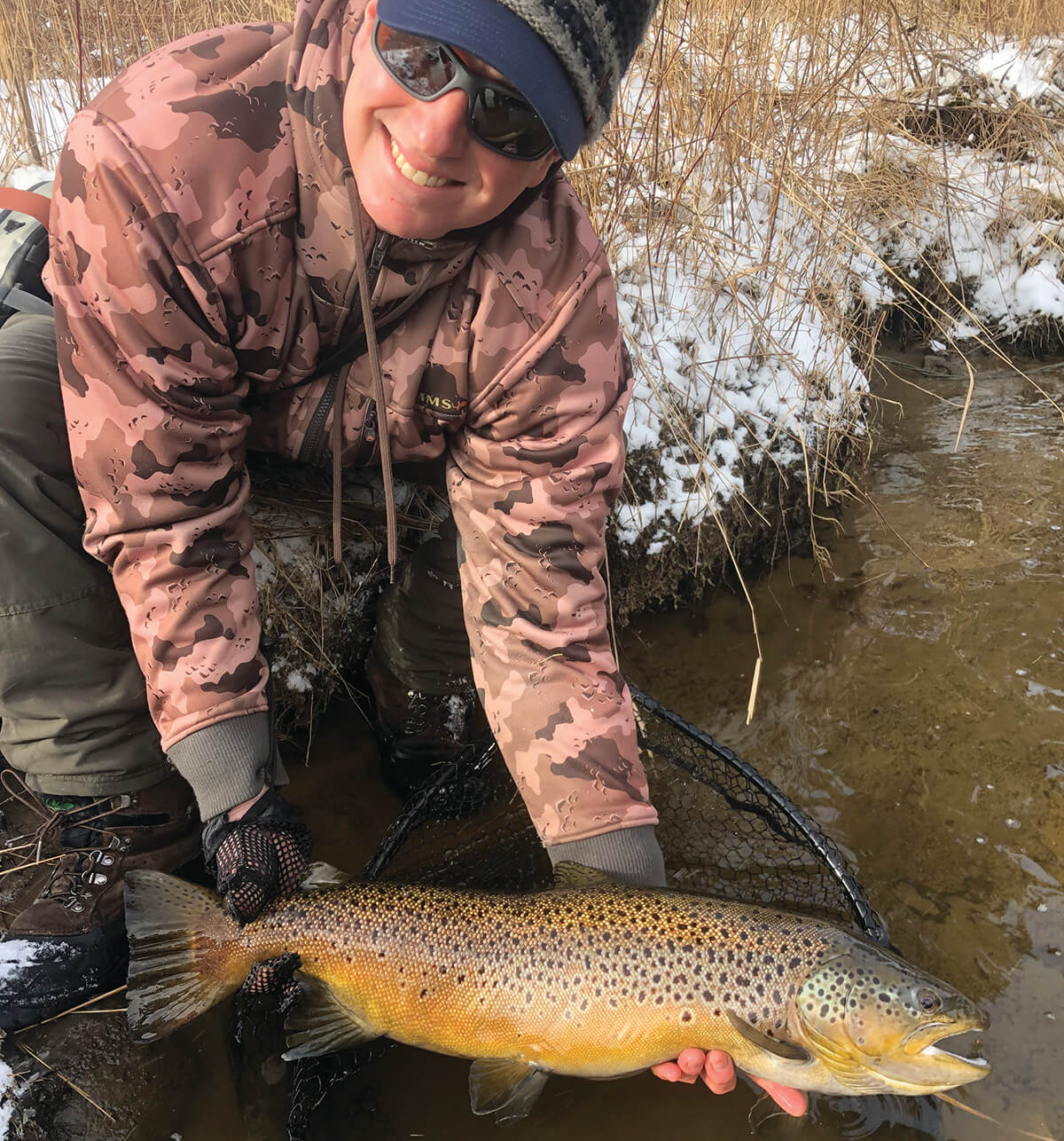 smiling angler holding large brown trout just above surface of water along a snowy bank