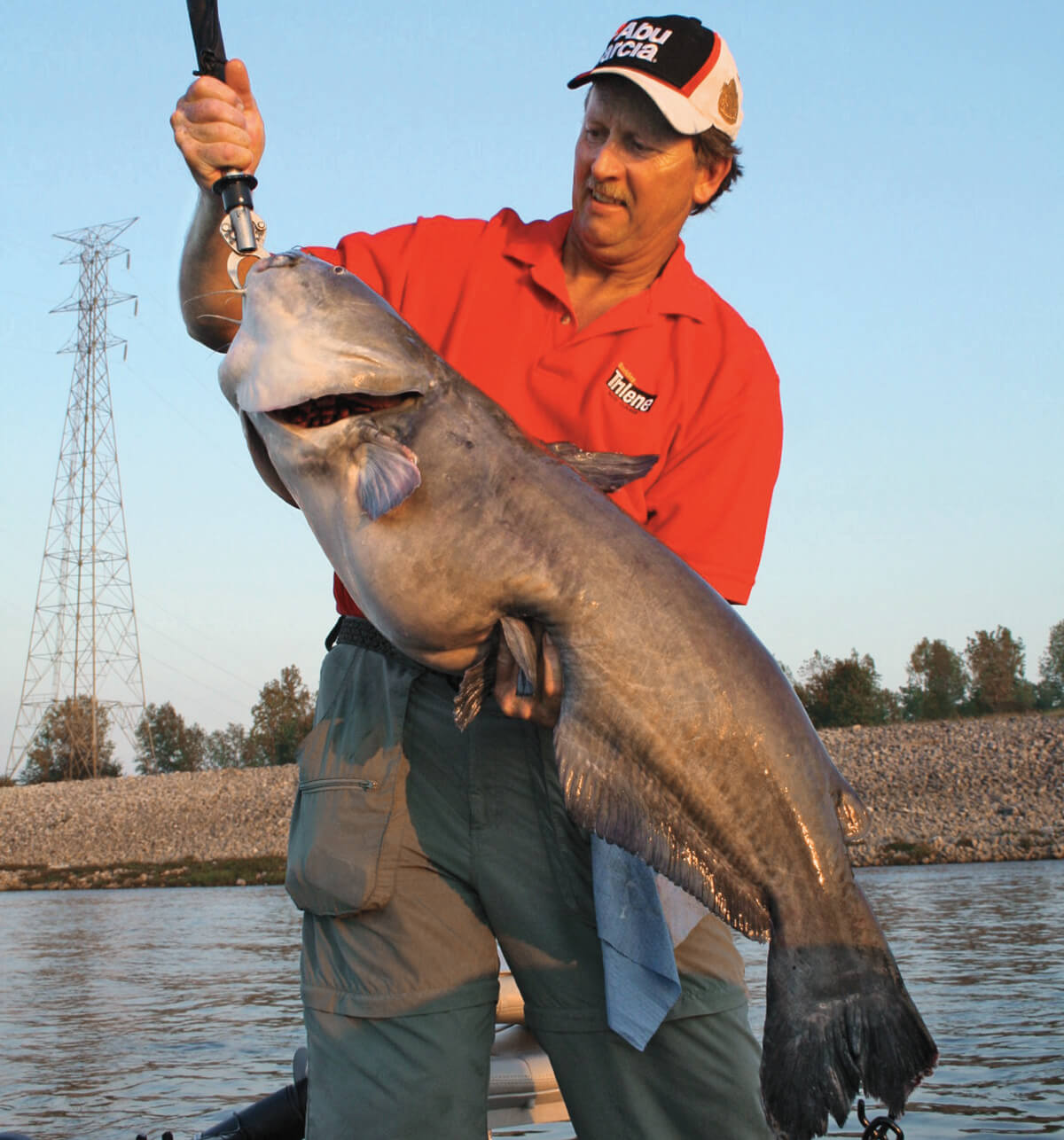 Angler holding a large catfish with a fish grip, in a boat on a river