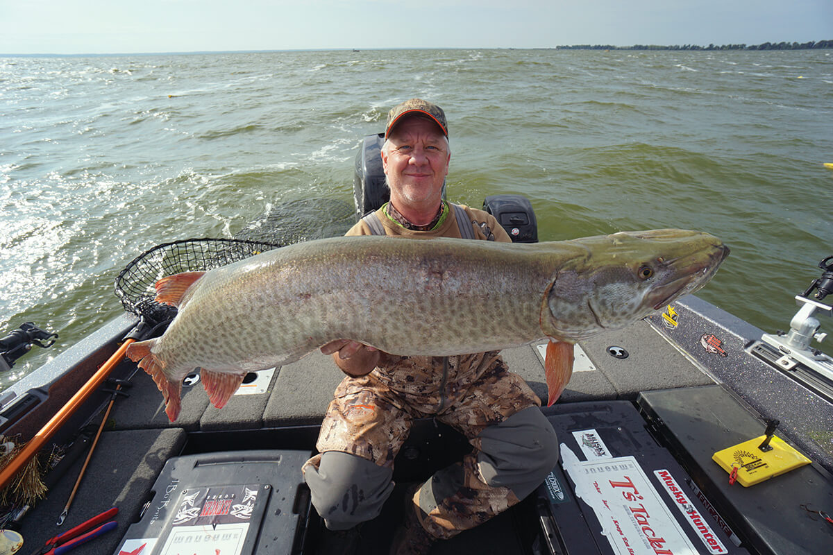 An angler holding a large muskie, sitting in the back of a boat on a lake