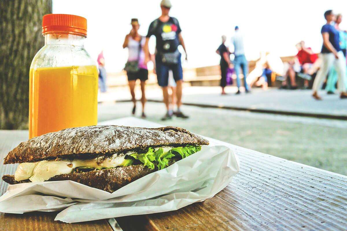 breakfast sandwich and bottle of orange juice on bench with tourists walking in background