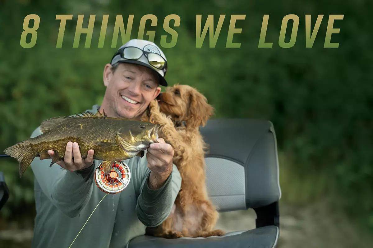 An angler smiling and holding a smallmouth bass while a puppy climbs on him.