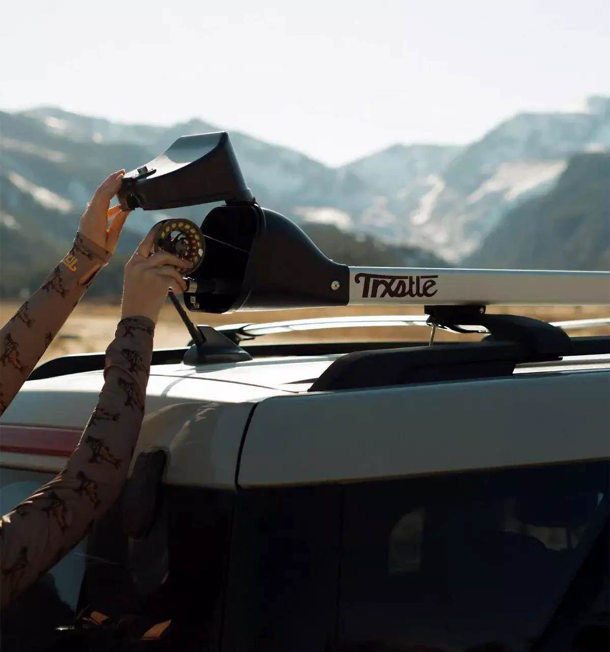 Two hands loading a fly rod into a Trxstle roof-top fly-rod carrier; mountains in the background.