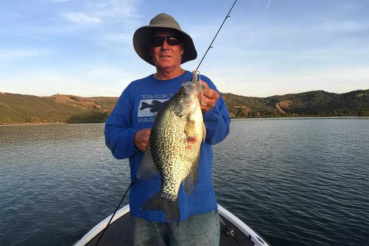 An angler with a fishing rod and reel in his hands poses with a large black crappie.