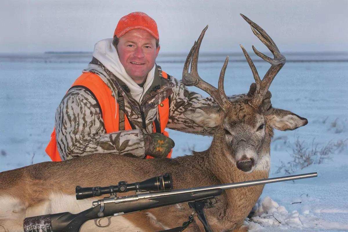 A hunter poses with a harvested white-tailed buck and his rifle in the snow. 