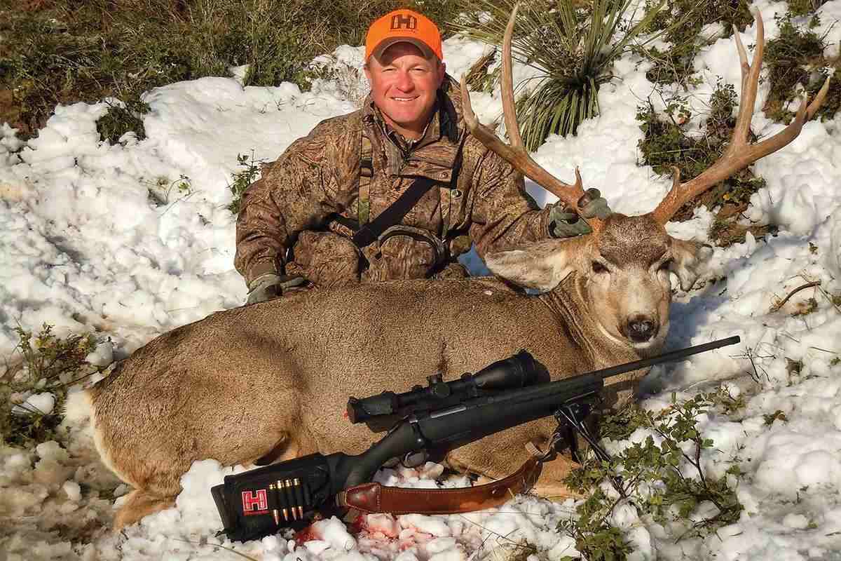 A hunter poses with a harvested mule deer buck and rifle. 