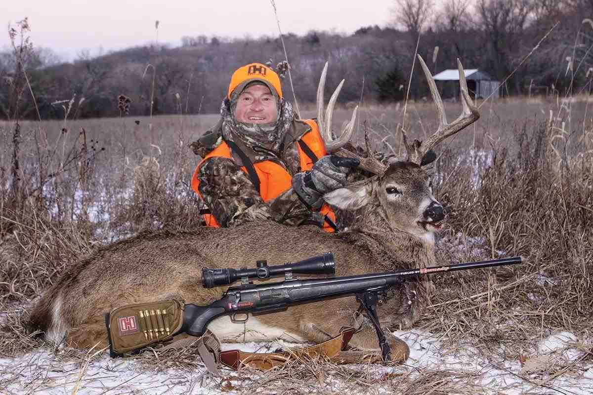 A hunter poses with a harvested buck with a rifle out front and a shed in the background. 