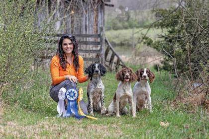 Amelia Baxter kneels next to her three springer spaniels with three ribbons next to them.