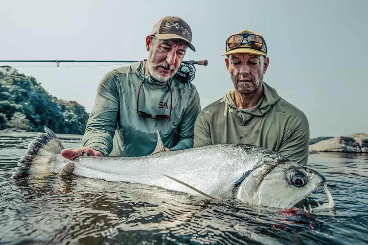 Two fly anglers together holding a sharp-toothed fish, a payara, wading waist deep with a fly rod on one's shoulders.