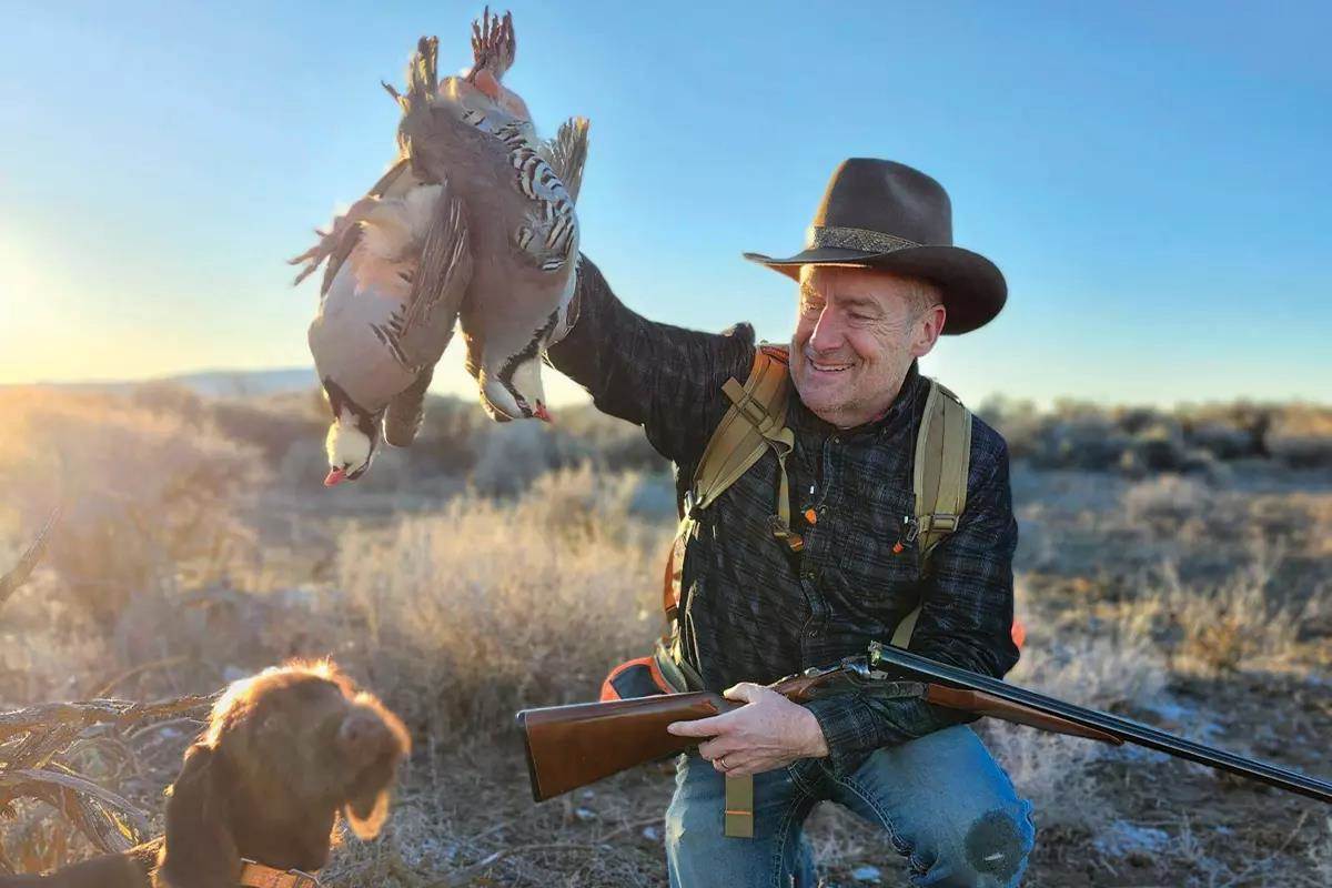 A hunter holds up two harvested chukar in front of his dog. 