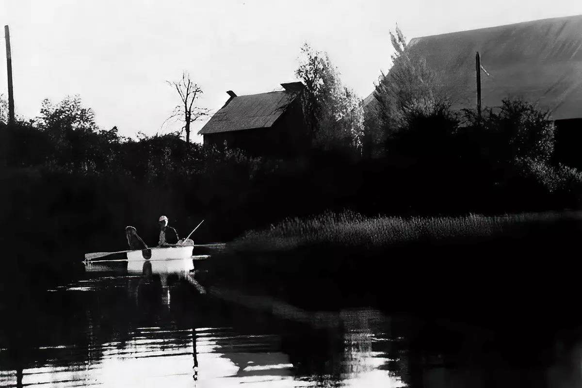 A black and white image of a man and a dog in a boat; barns in the background.