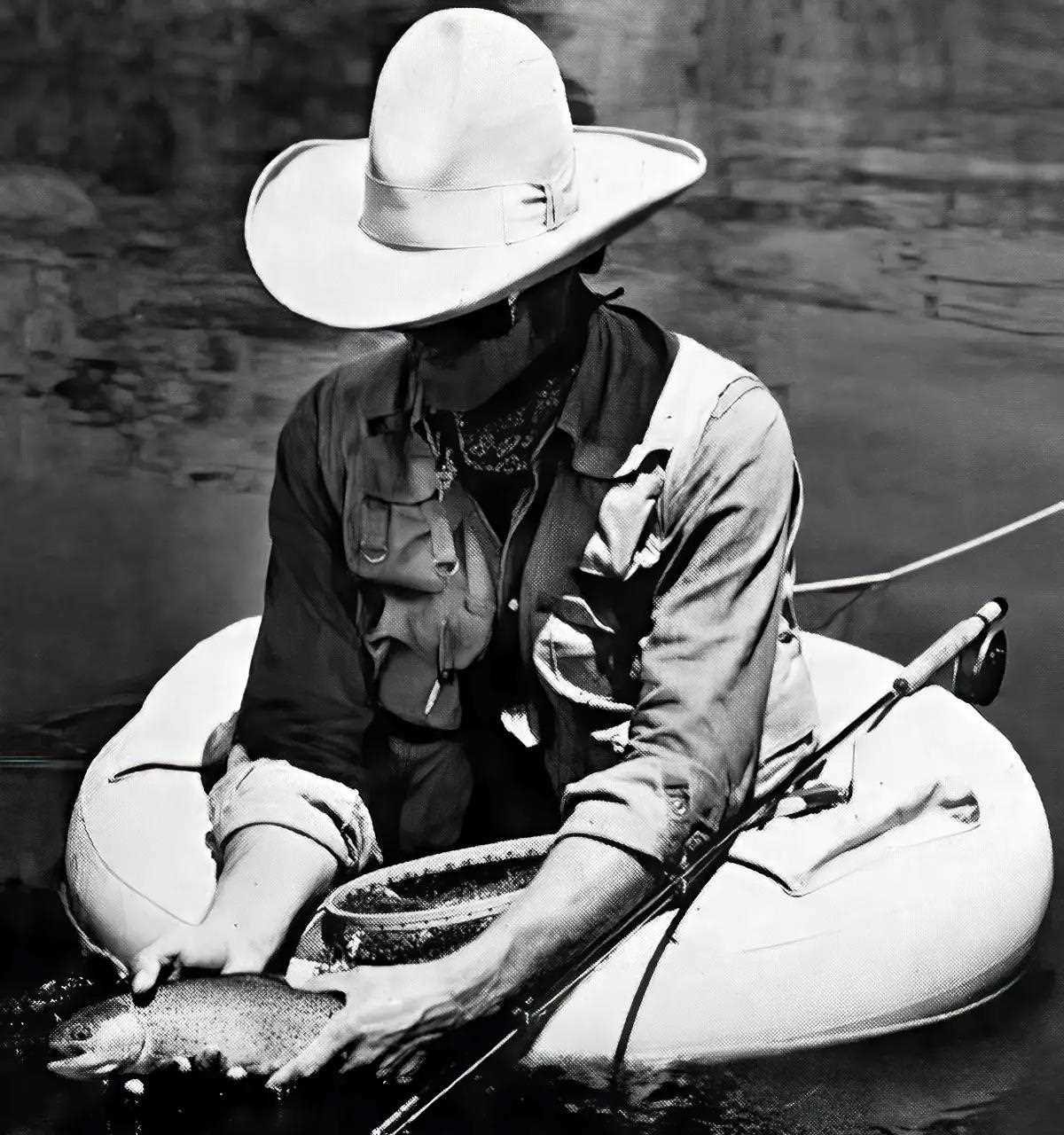 A black and white photo of a fly angler in a cowboy hat holding a trout in a float tube.