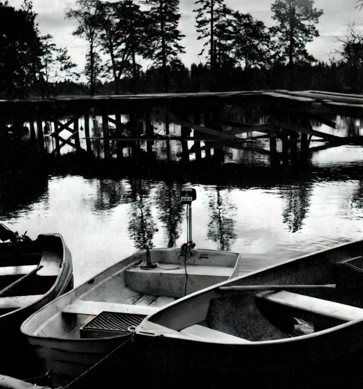 A black and white photo of several boats in the foreground and a wood bridge in the background.