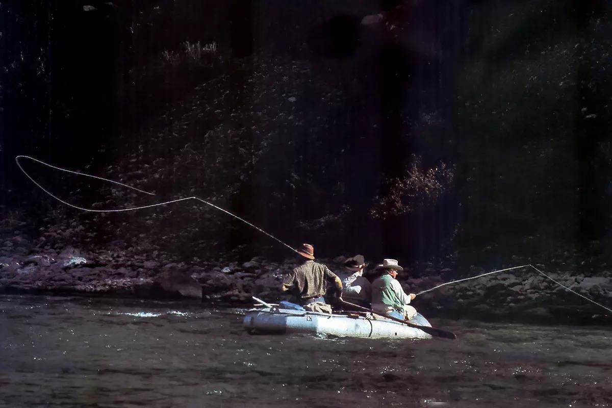 Three people in a raft on a river; one is rowing, two are fly fishing. 