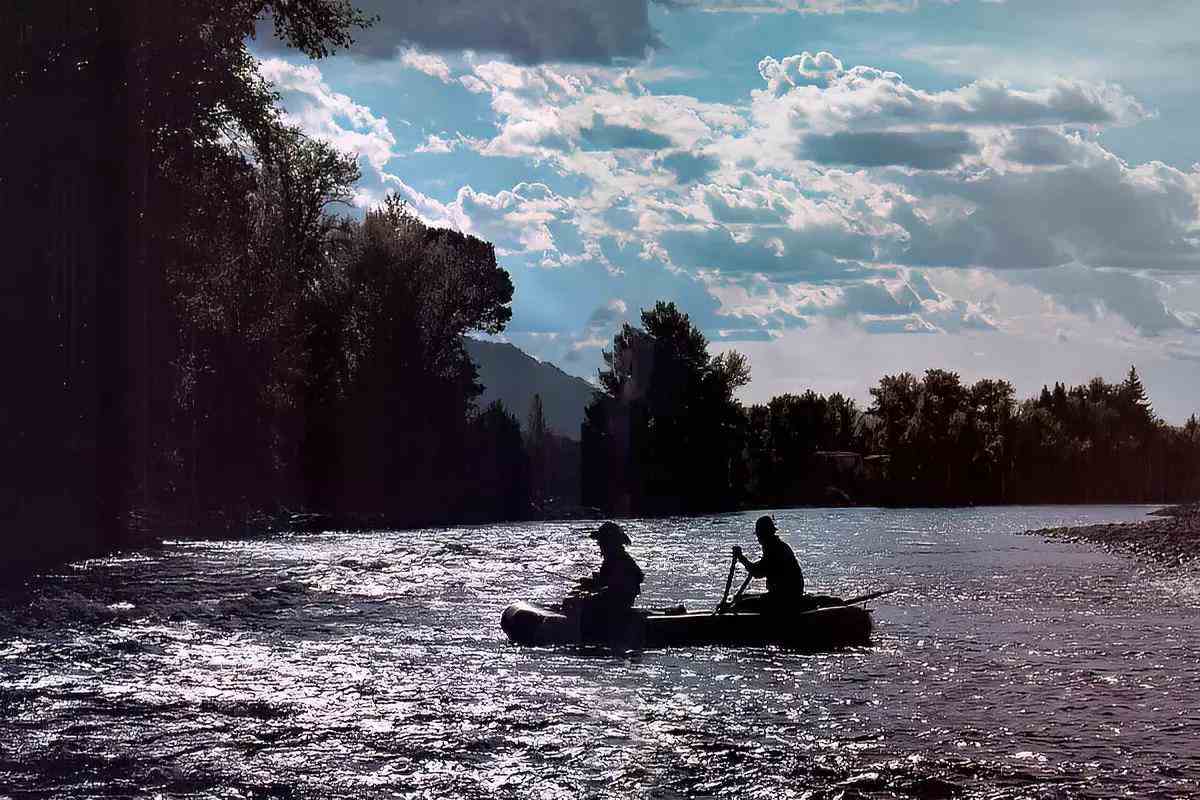 Two fly anglers in a boat silhouetted against the sheen of a river. 