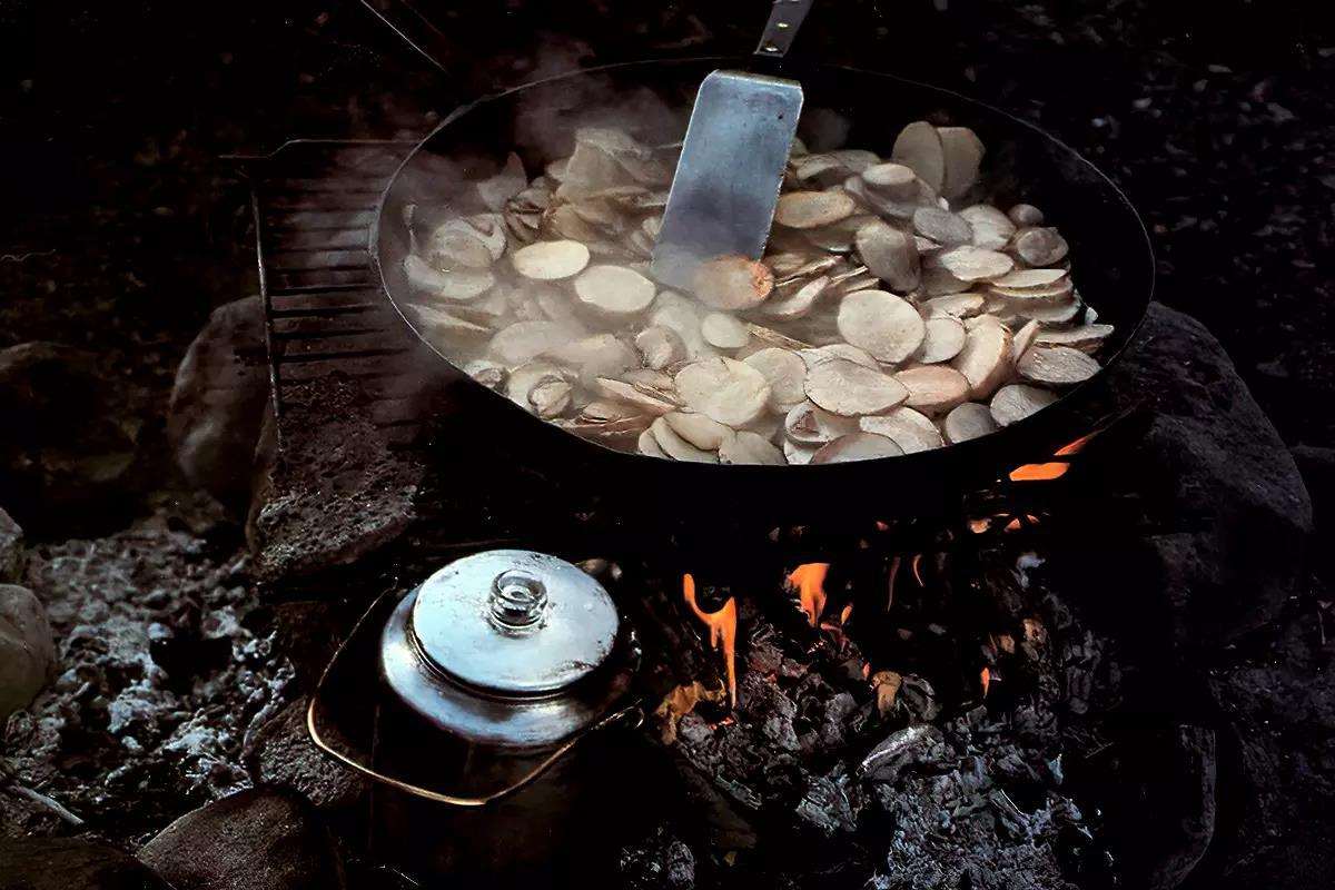 Sliced potatoes frying in a cast-iron pan over a campfire; a percolator coffee pot in the foreground.