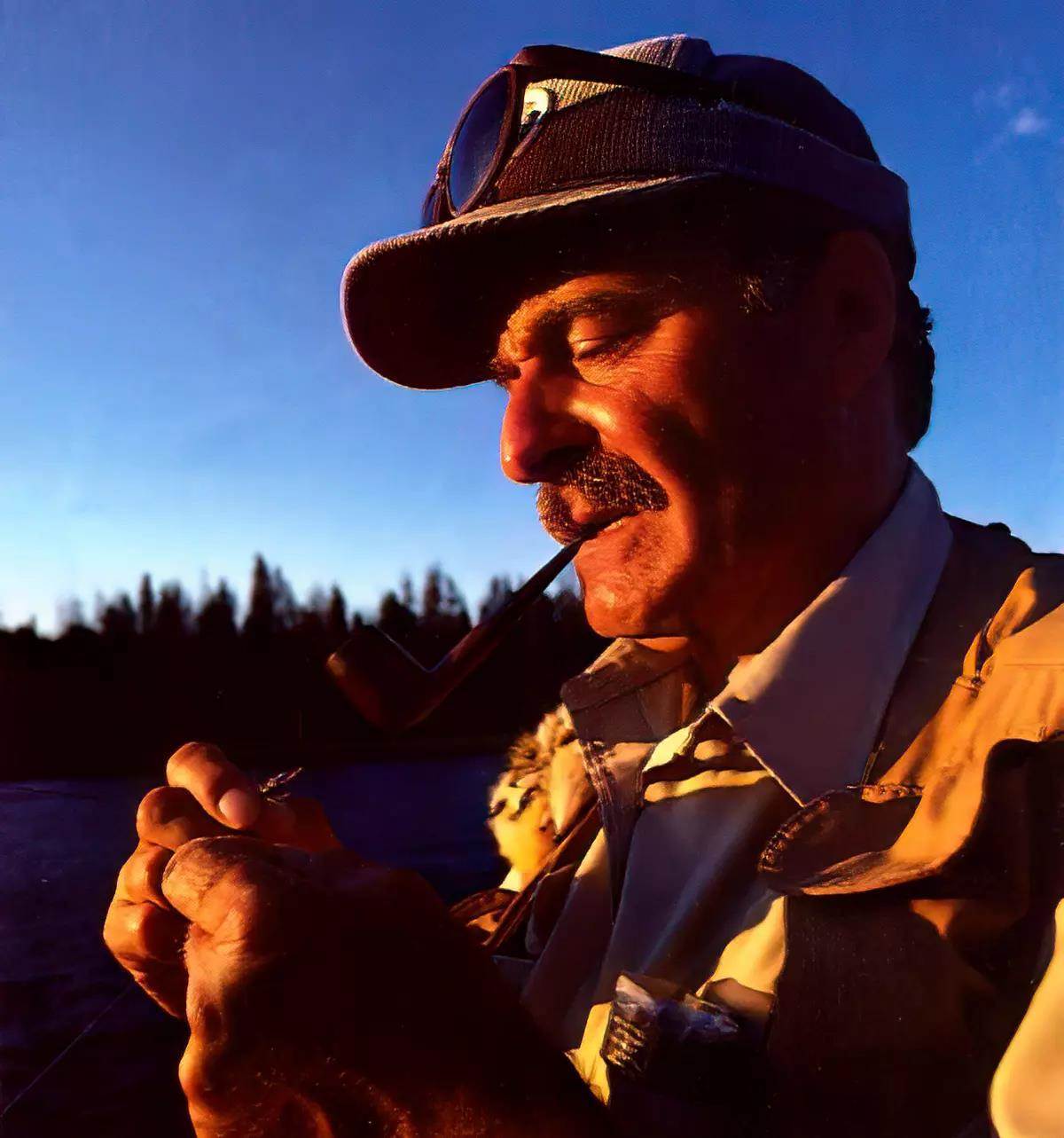 A fly angler with a pipe in his mouth tying on a fly. 