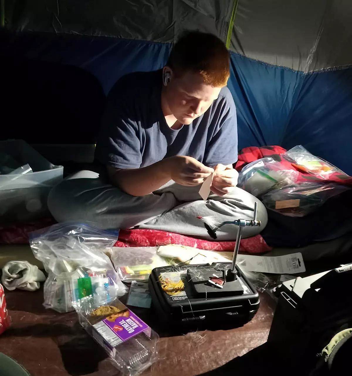 A teenaged boy tying flies inside a tent. 