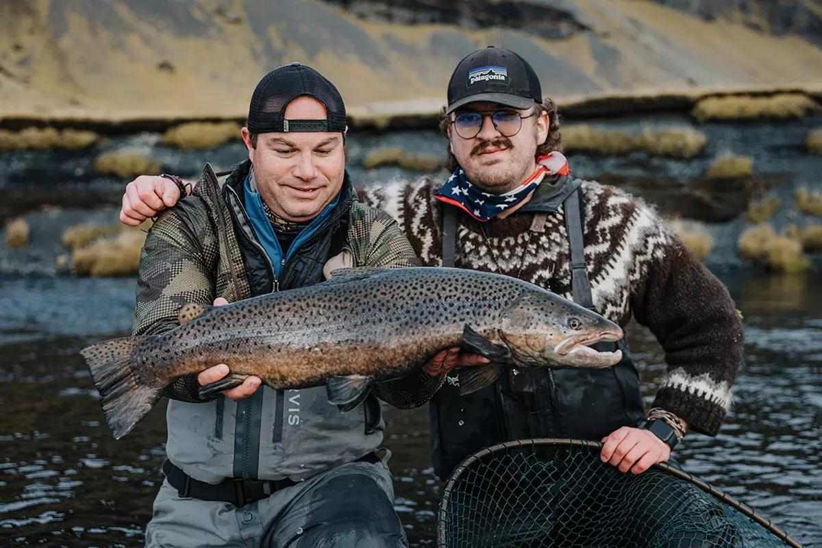 Two fly anglers posing arm-in-arm for the camera, one holding a large brown trout. 