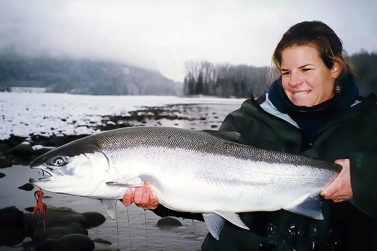 A woman holding a large chrome steelhead with a fly hanging from its mouth on an overcast day