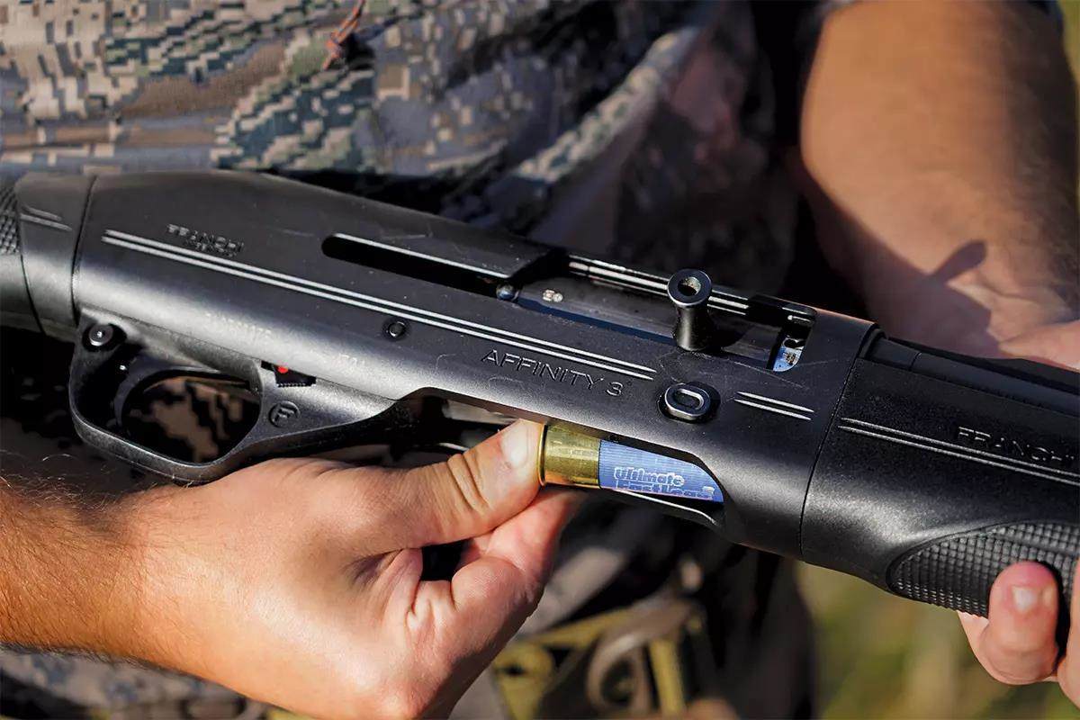 A hunter loads a blue shotgun shell into a black shotgun.
