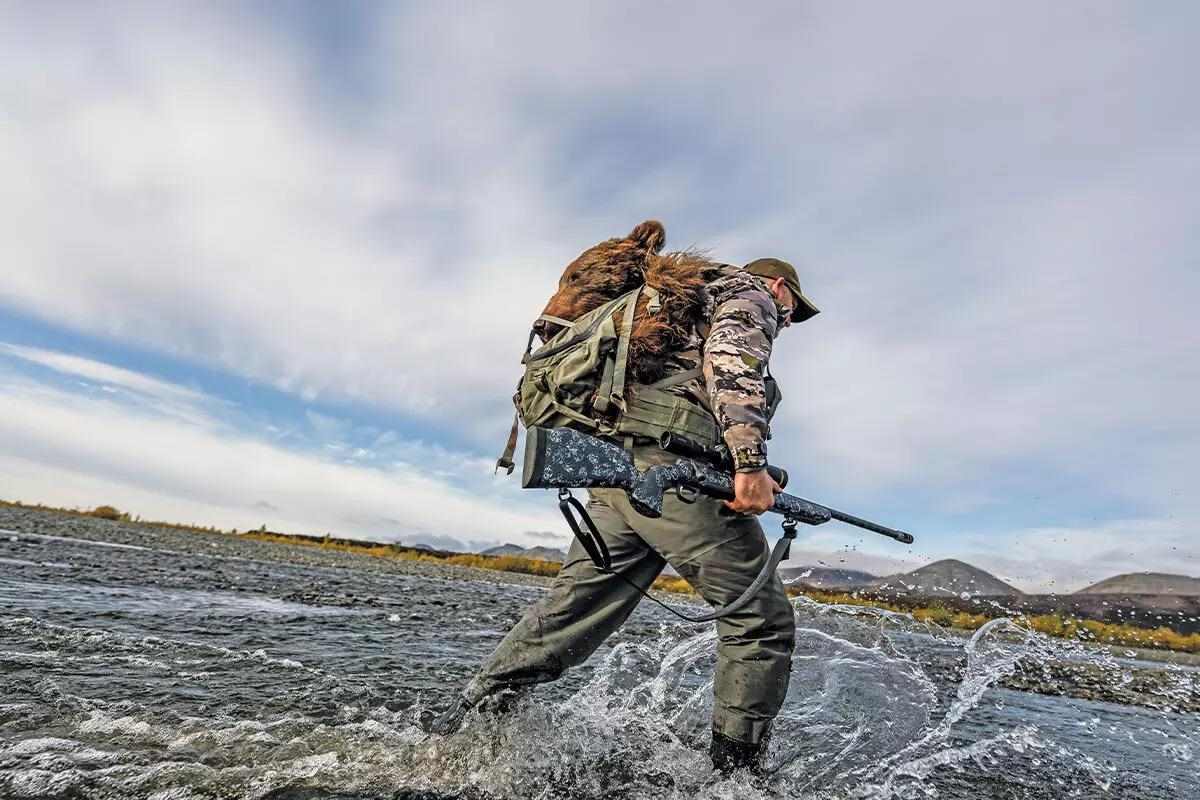 man tromping through creek water with rifle and backpack