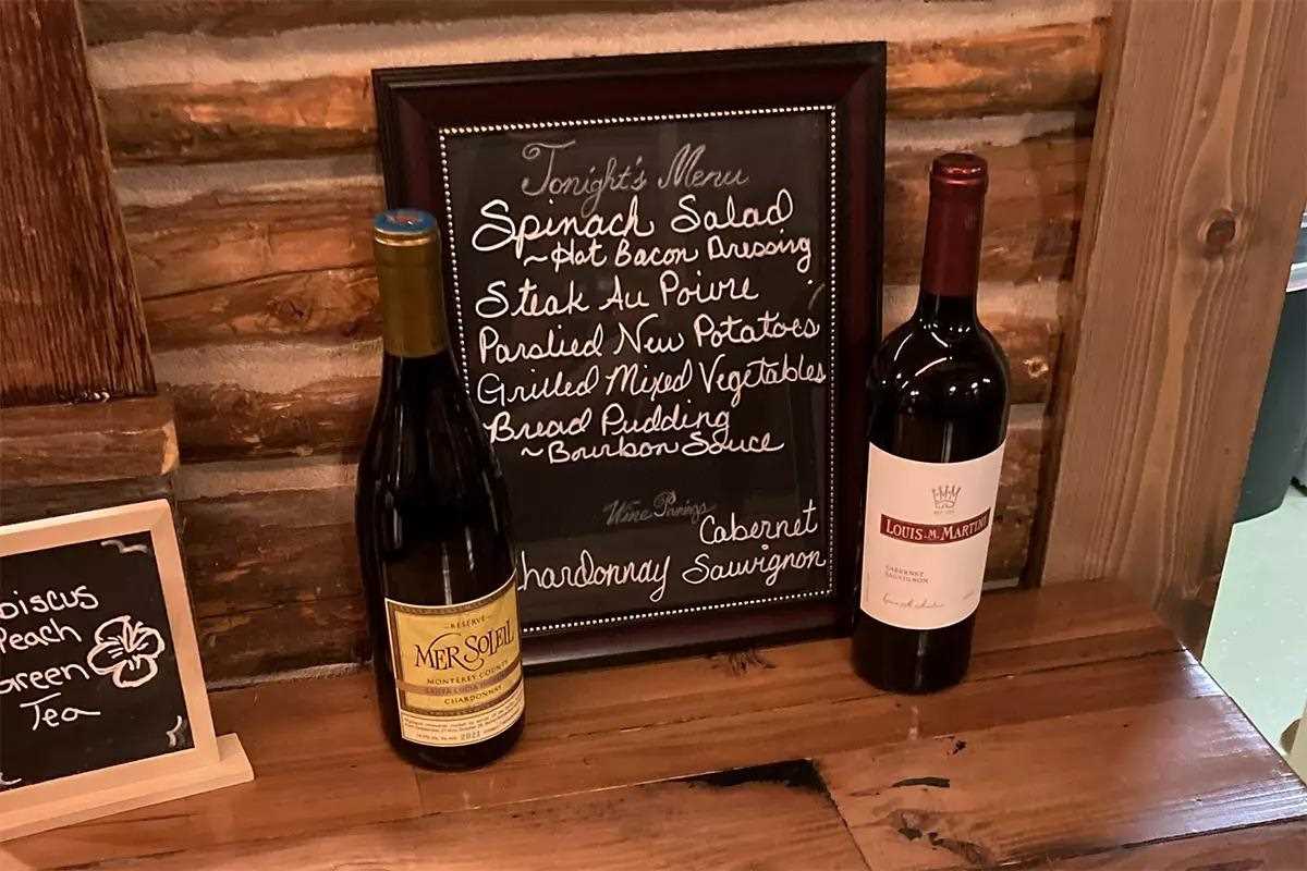 A chalkboard menu is displayed on a wooden shelf with items like spinach salad, steak au poivre, and bread pudding. Two bottles of wine, Mer Soleil and Louis M. Martini, are placed next to the menu.