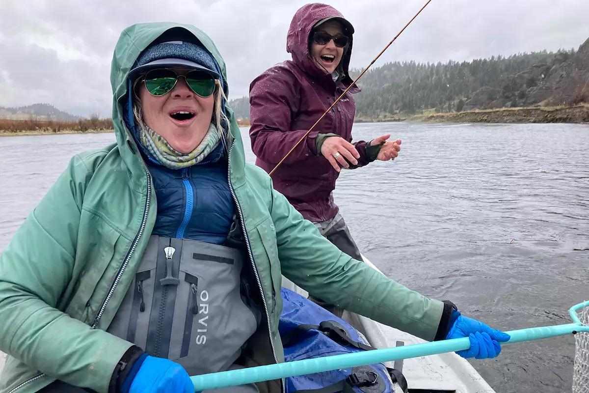 Two people are in a boat on a river. They are wearing rain jackets and gloves, and one is holding a fishing rod while the other holds a net labeled 