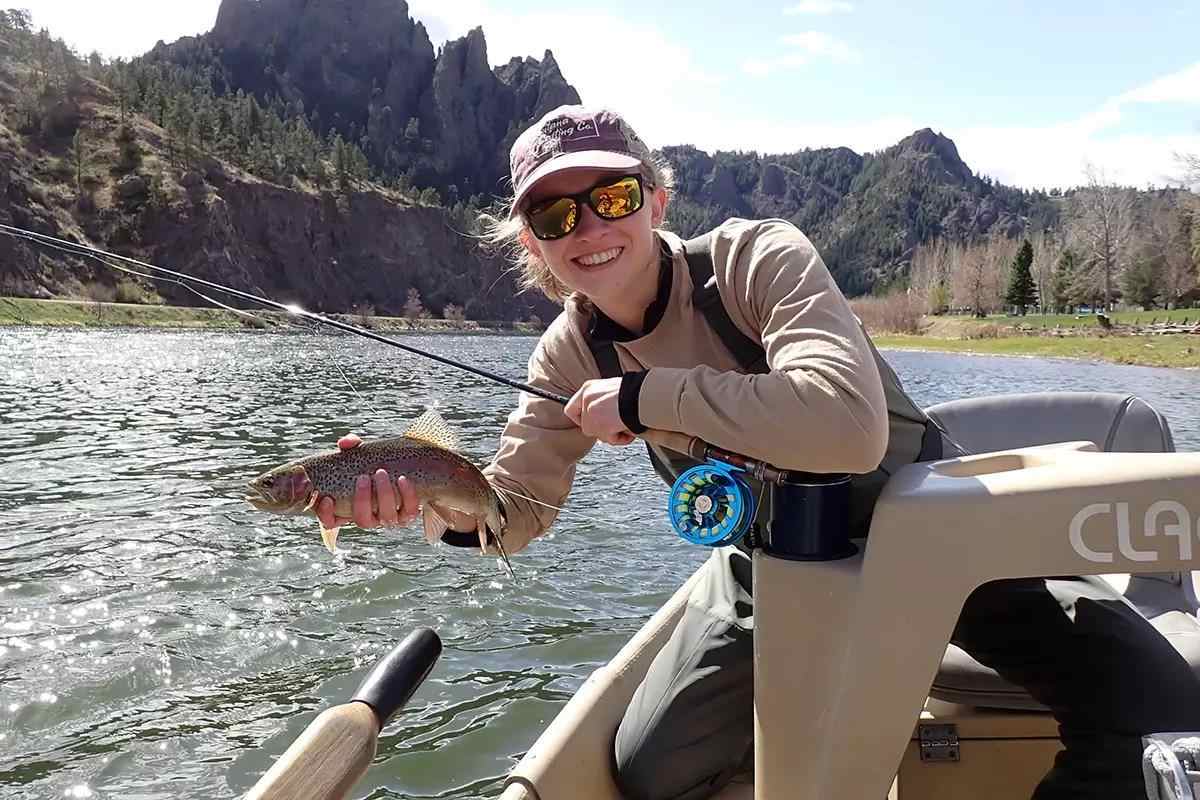 A person smiling while holding a fish on a fishing rod in a small boat. The location is a scenic river with mountainous terrain and sparse trees in the background. The person is wearing sunglasses, a hat, and waders.