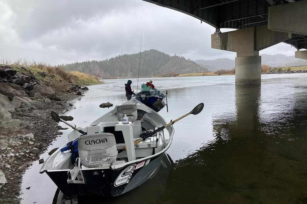Two drift boats are positioned on a calm river under a large concrete bridge. The foreground boat is labeled 
