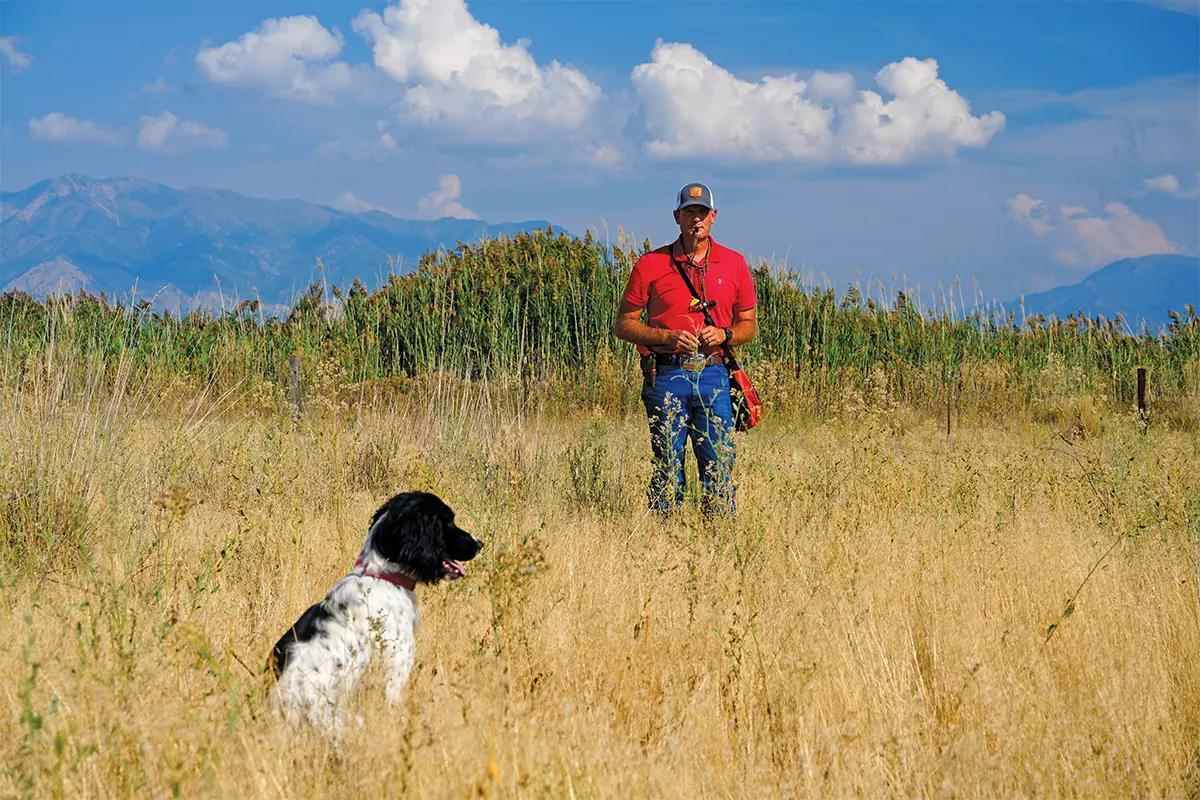 A dog trainer stands with a whistle in his mouth while a black and white dog sits in the field in front of him.