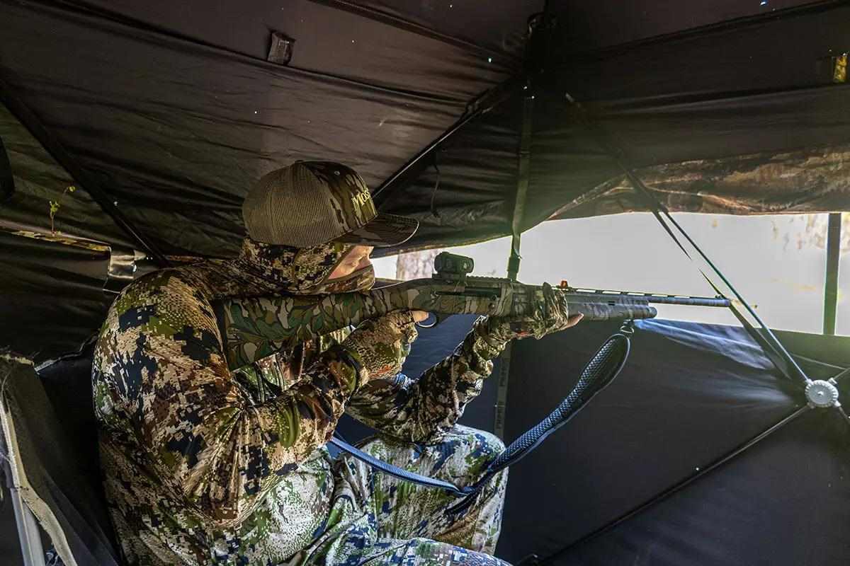 Turkey hunter sitting in ground blind with shotgun at the ready.