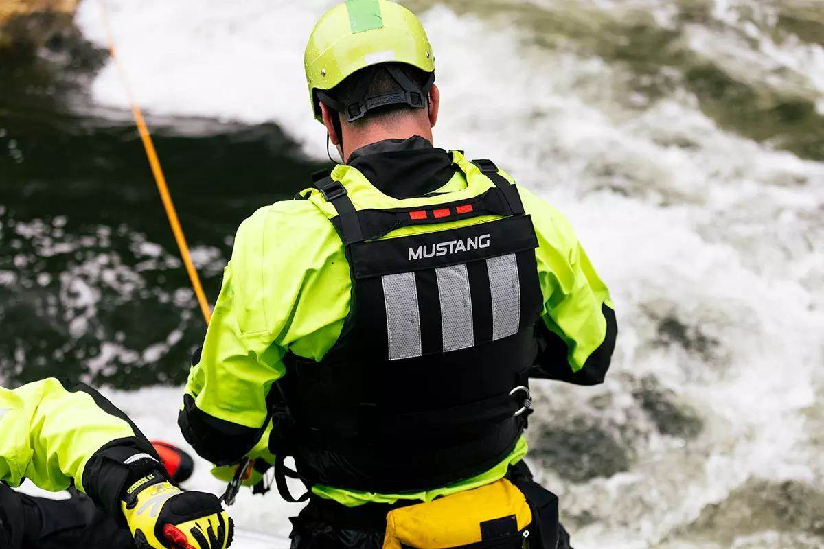 A river rescue professional wearing bright green, a helmet, and PFD next to a whitewater section of river.
