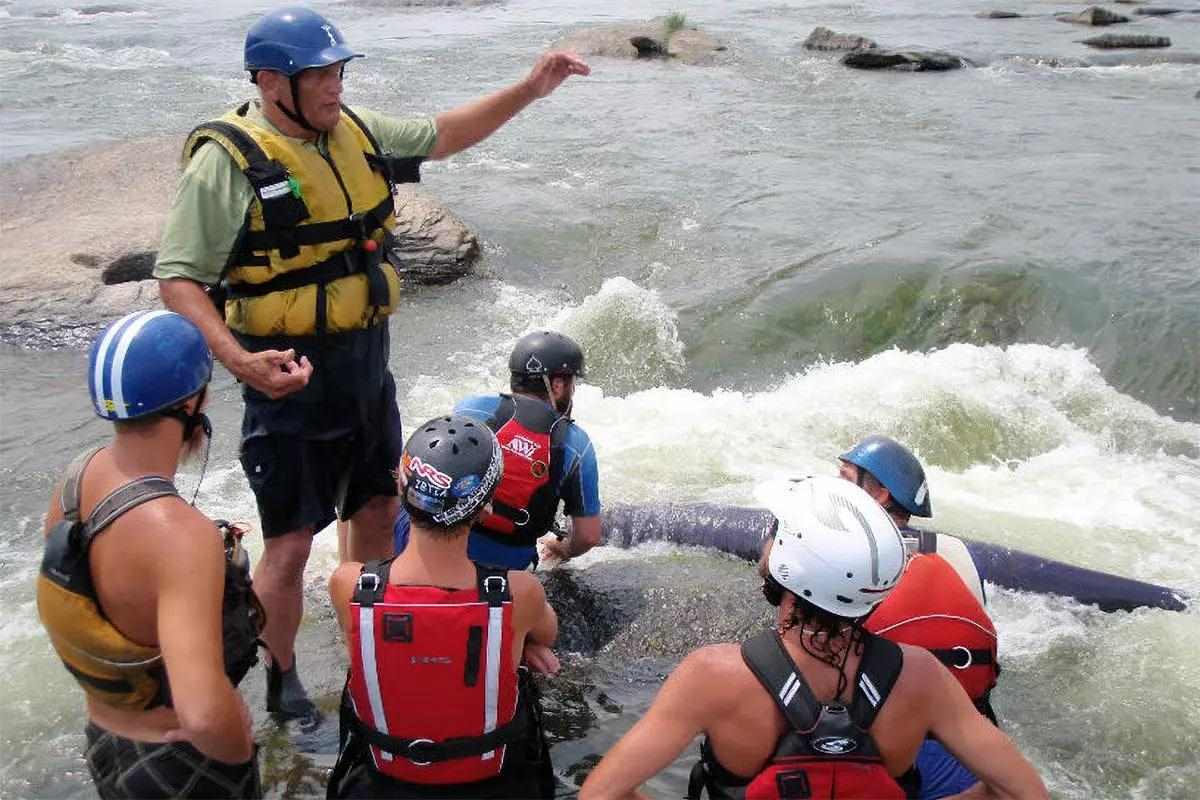 A man wearing a helmet and PFD talking to a group of others also wearing helmets and PFDs on a swift section of a river.
