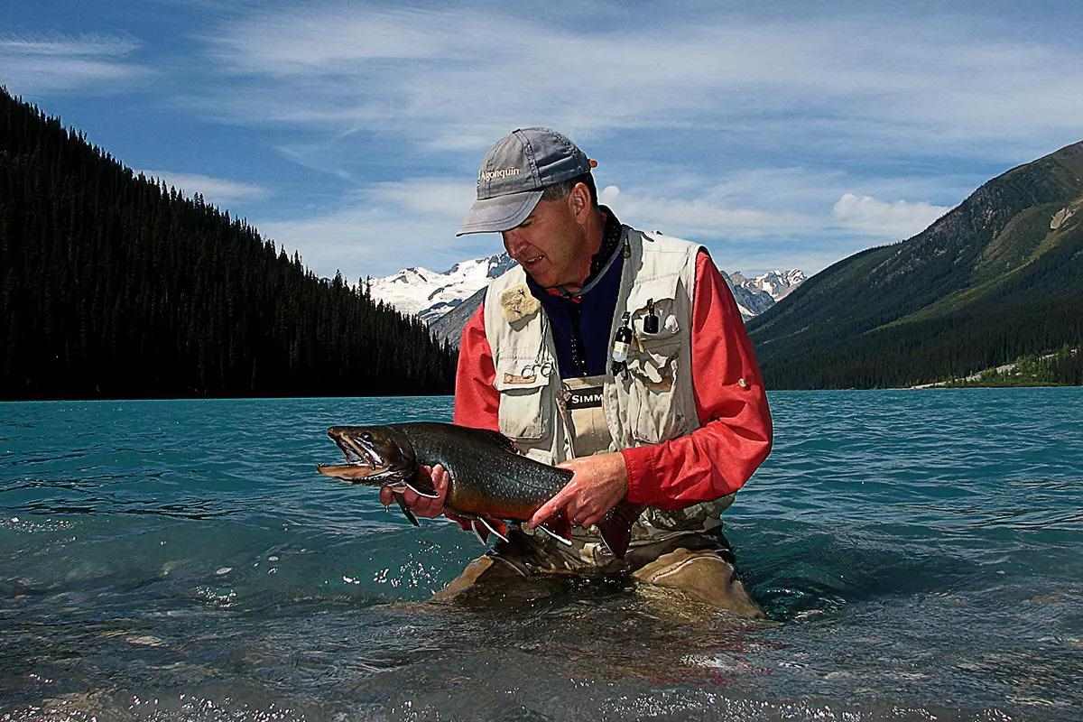 A man kneeling in the shallow water of a lake, holding and looking at a large brook trout.