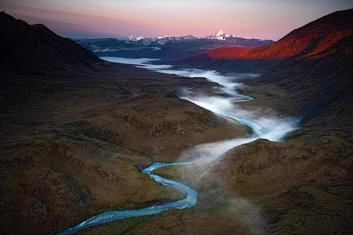 A landscape photo of a foggy river exiting a lake at dawn, beneath large mountains.