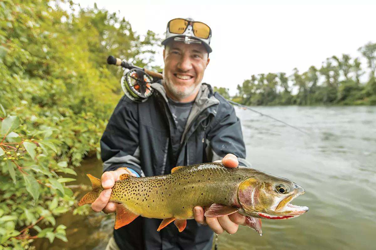 A smiling man holds a cutthroat trout; a fly rod and reel over his shoulder.