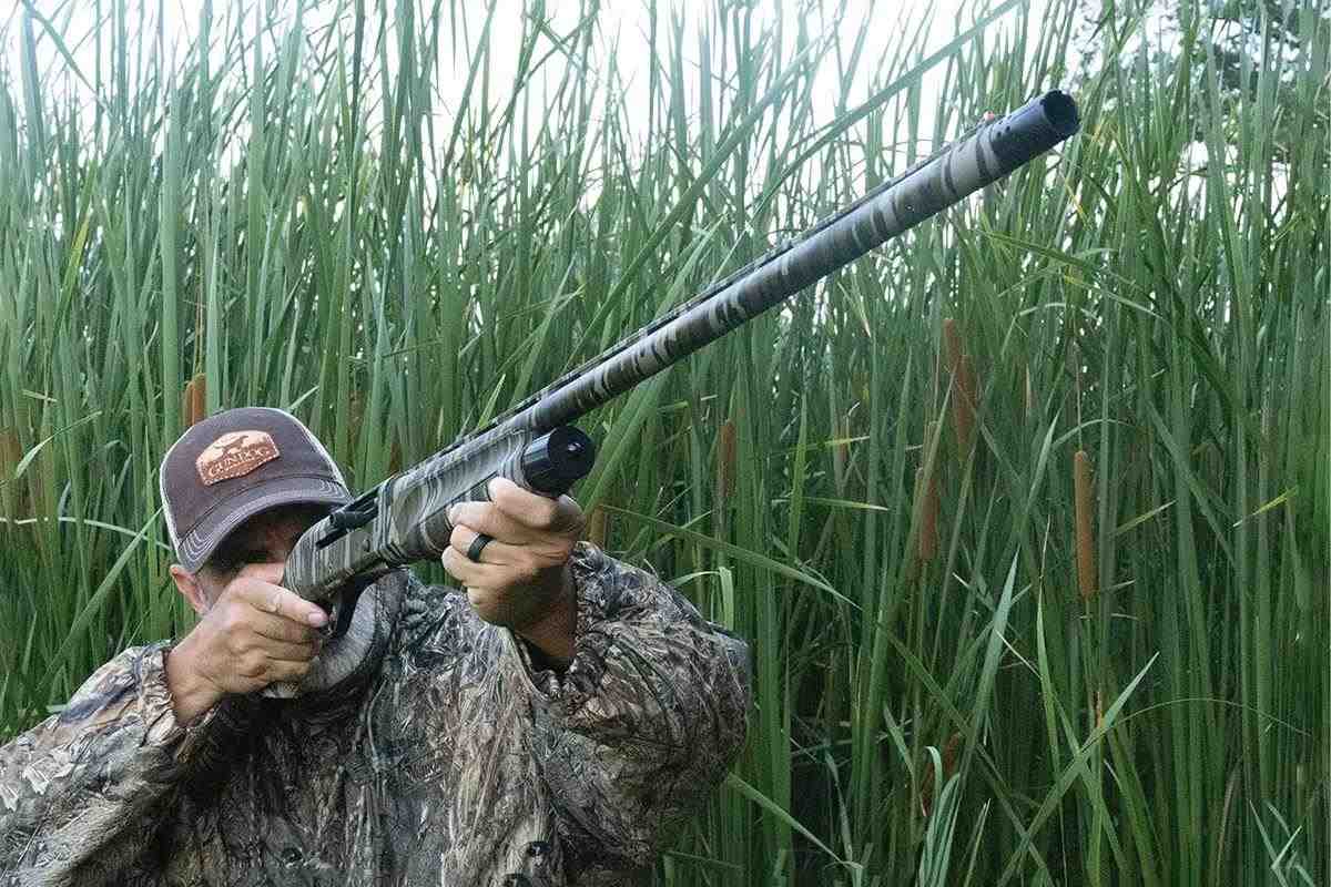 A hunter wearing camo in tall reeds aims down the barrel of a camo shotgun.