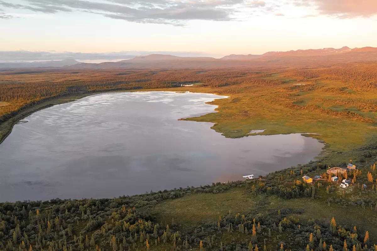 An aerial photo of a lodge being built next to a lake in a remote area. Mountains in the distance.