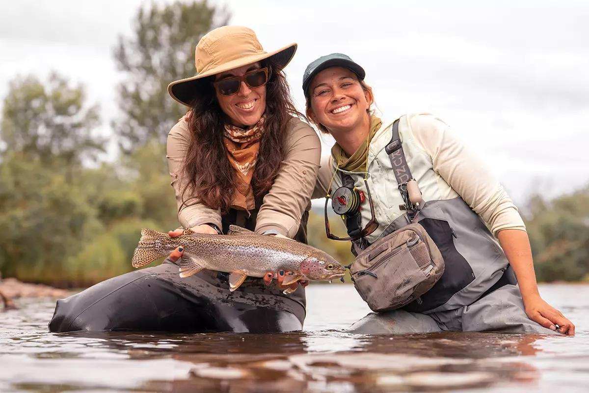 Two fly anglers kneeling in a river, one holding a rainbow trout.