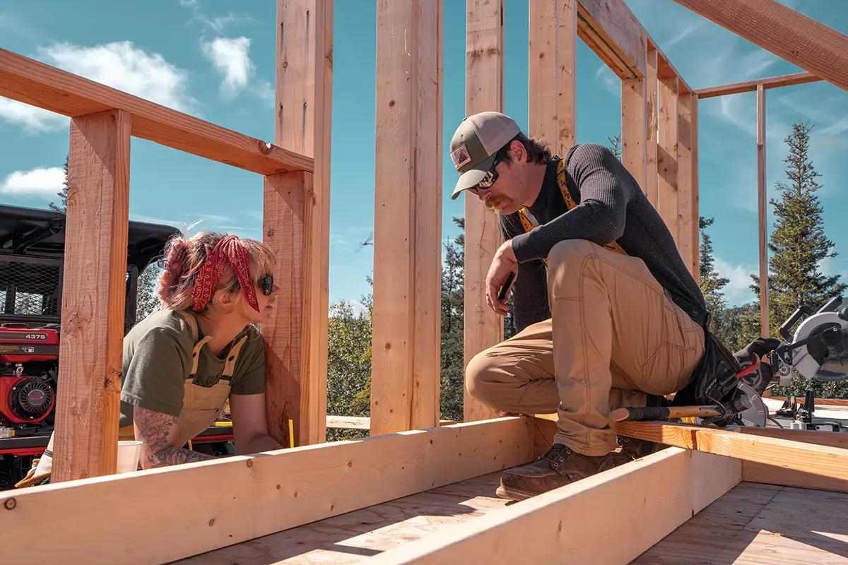 A man talking to a woman through some studs on a framed house in progress.