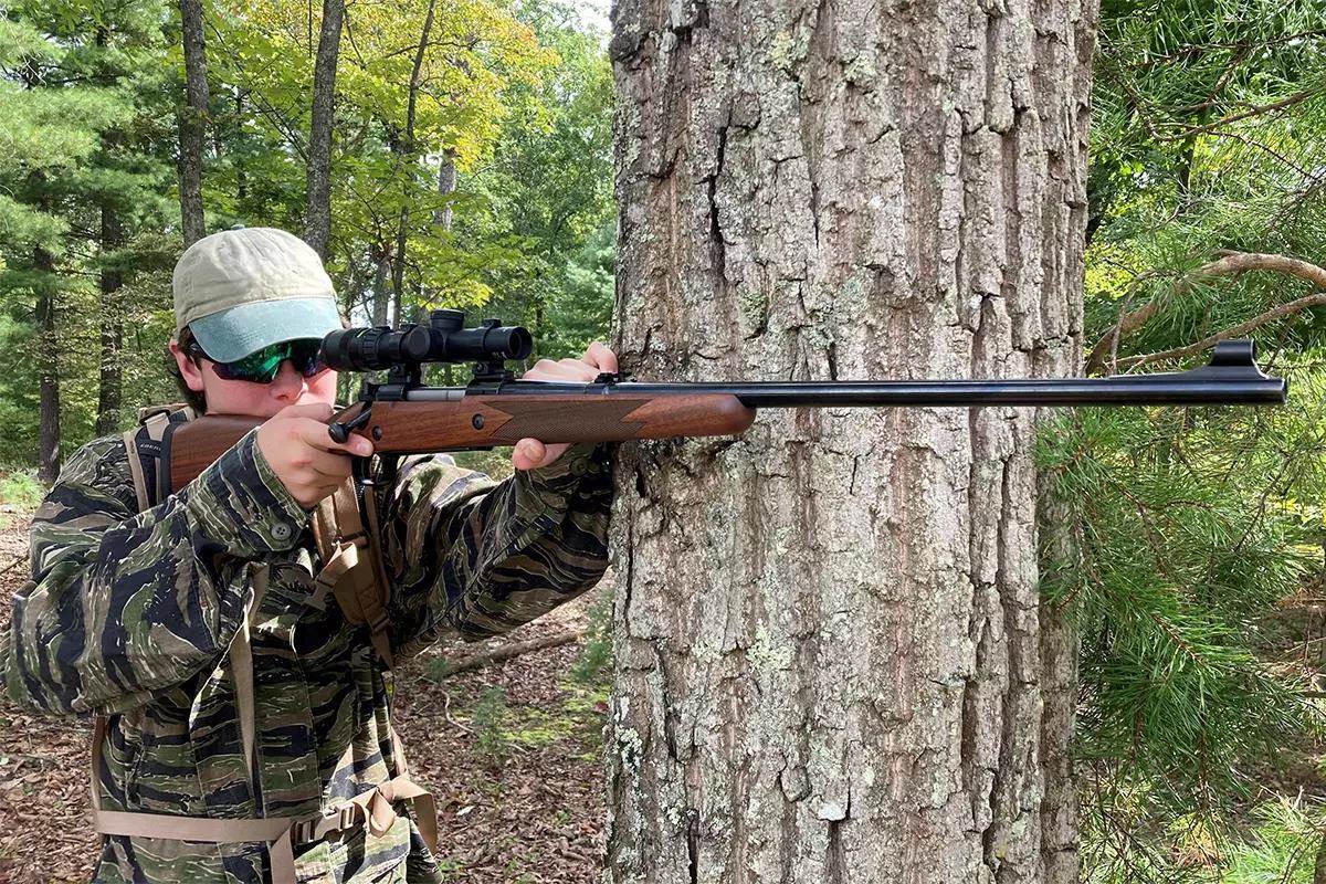 A shooter leans a rifle against a tree while aiming the rifle. 