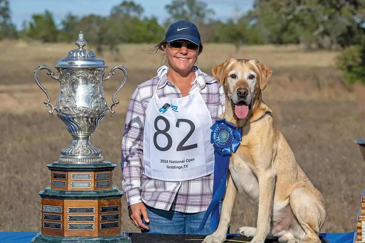 Just Floyd sits with his owner and a giant silver trophy after winning the 2024 National open retriever field trial.