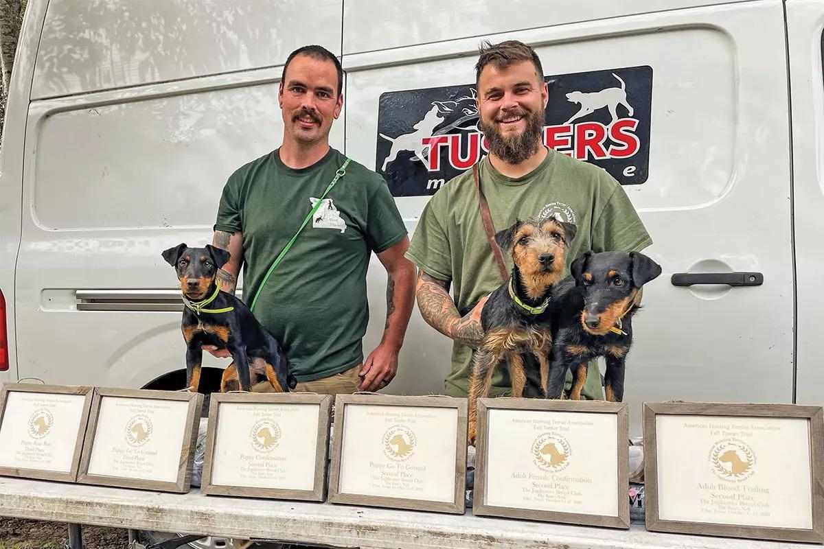 Two guys stand with three terrier dogs and a bunch of award certificates.