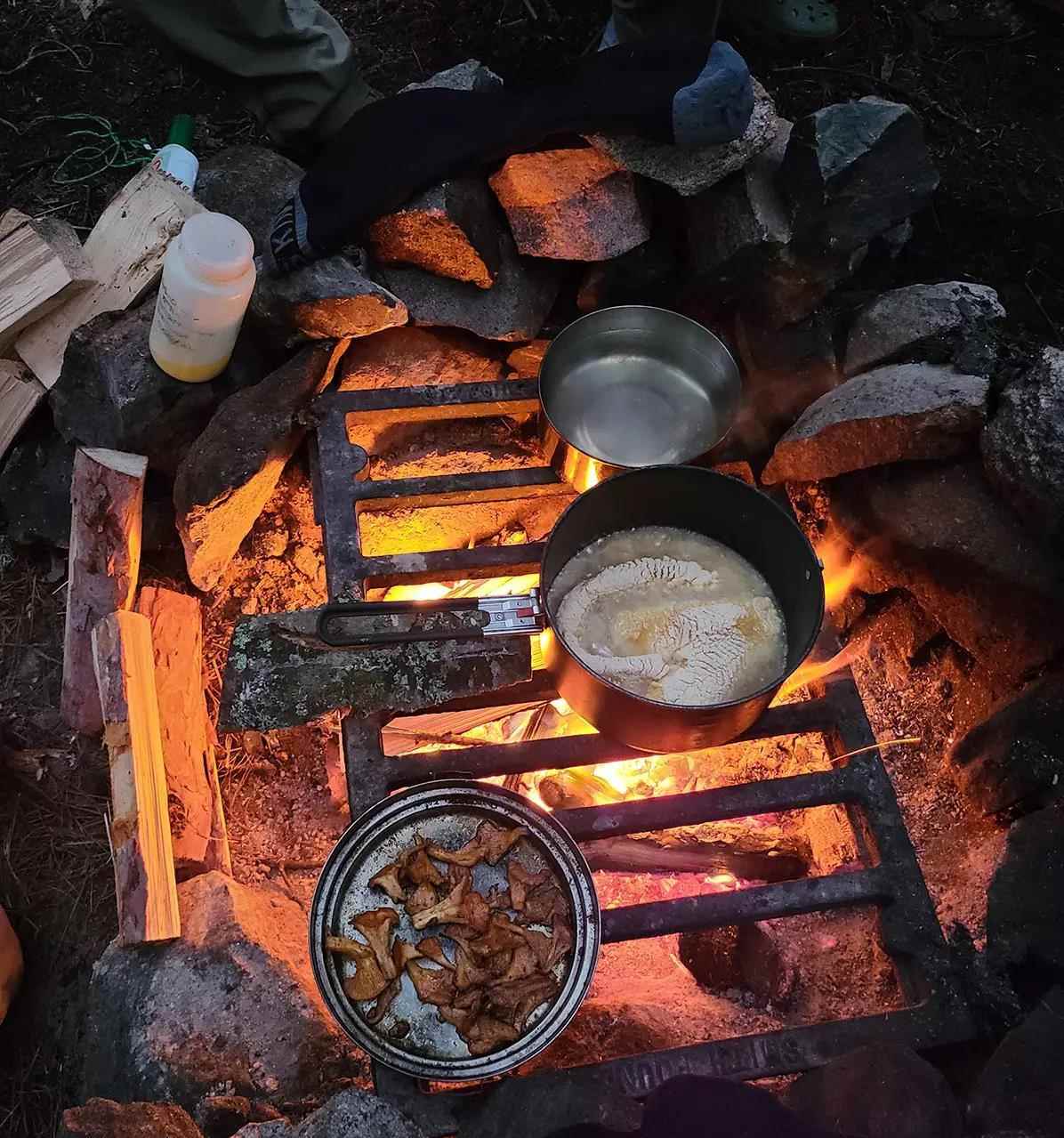 A grill grate over a rock-lined campfire with pots of frying fish cooking.