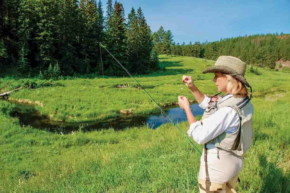 A fly angler wearing hip waders adds a strike indicator to her tippet, standing in green grass next to a small creek. 