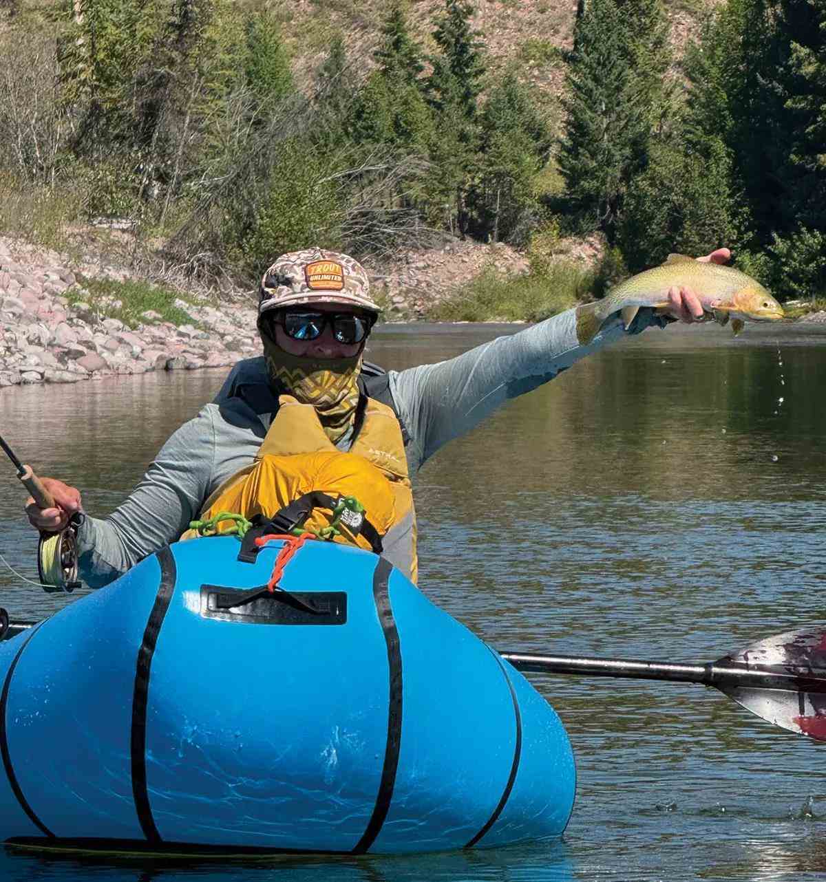 A fly angler in a small blue pack raft holding up a cutthroat trout.