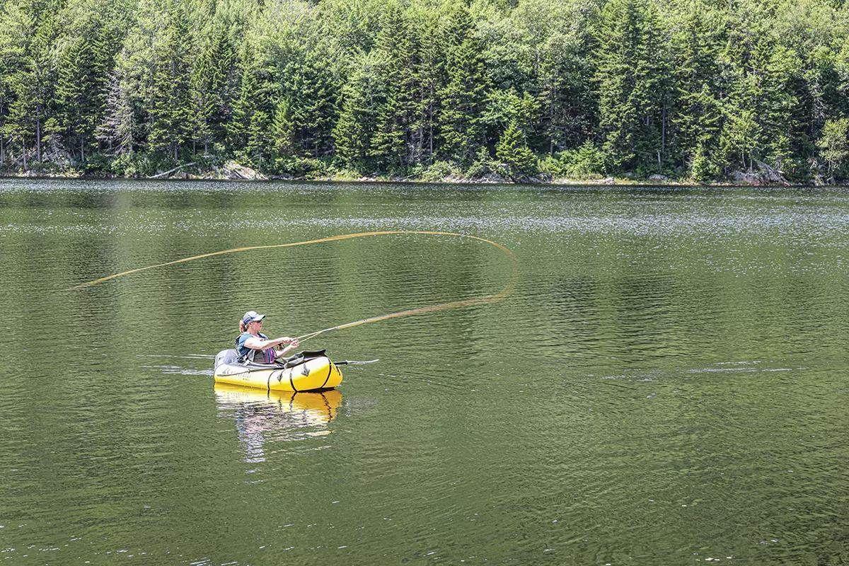 A fly angler in a small yellow raft casting on an pine-tree-lined lake.