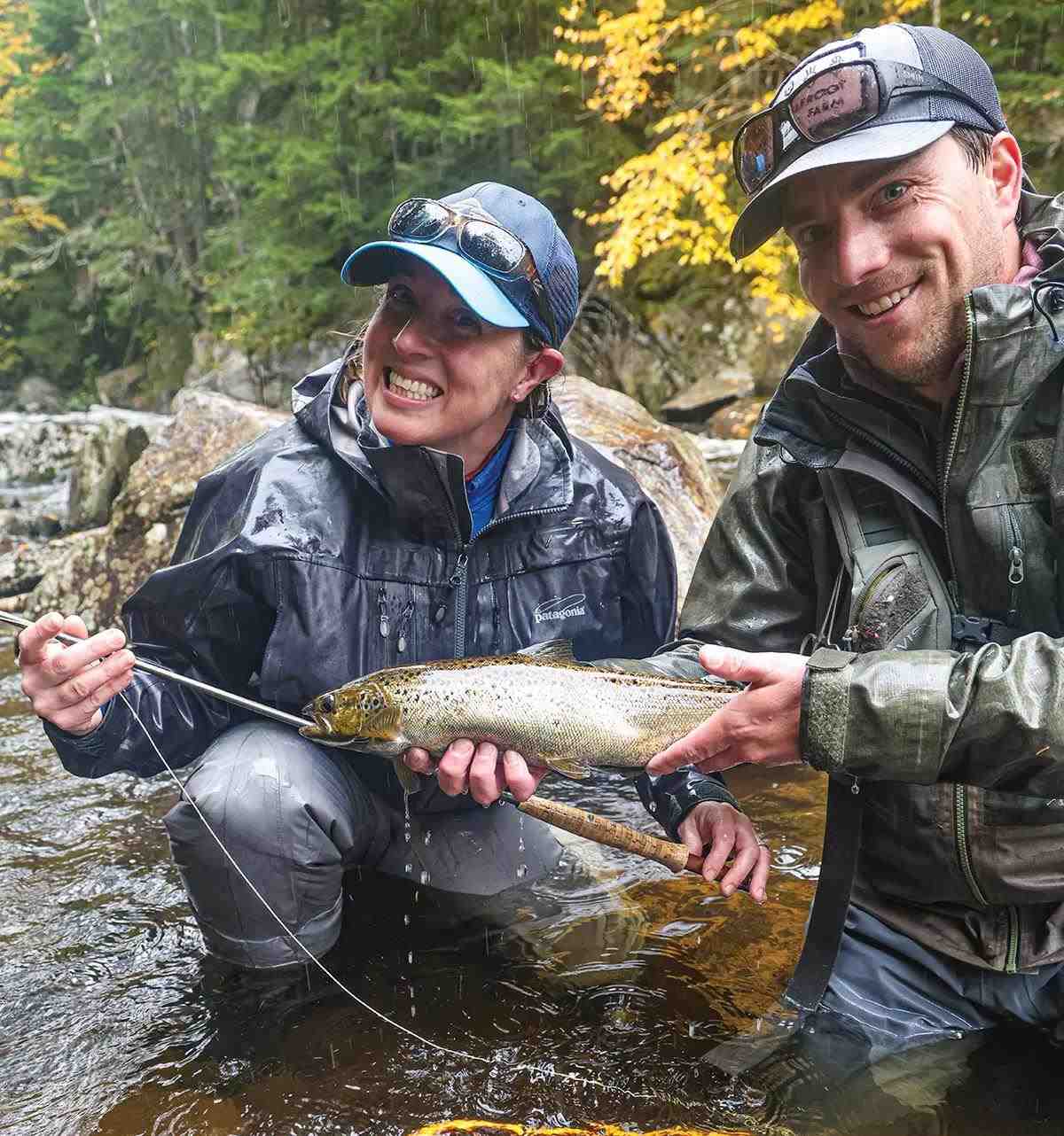 Two fly anglers squatting in a stream; one is holding a brown trout, the other is smiling holding a fly rod.