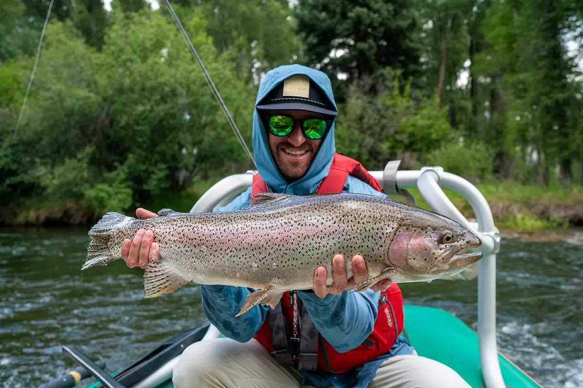 A fly angler in a raft holding a large rainbow trout. 